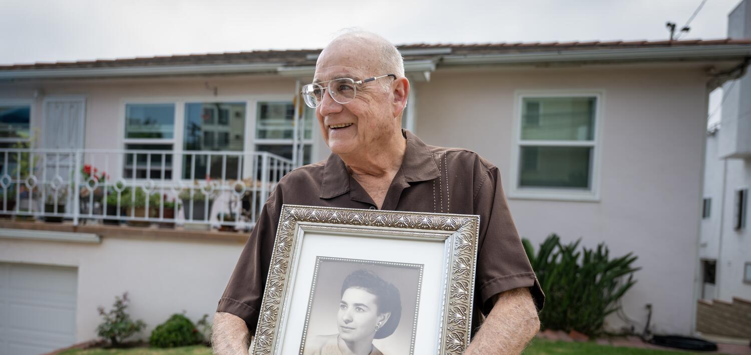 An elderly man, holding a black-and-white photo of his mother, stands in front of his Little Italy home.