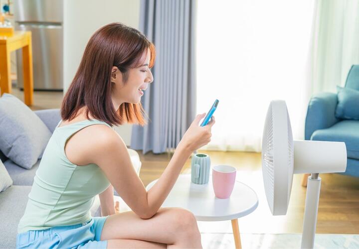 A woman sits on her couch, looking at her mobile phone with a fan placed in front of her.  