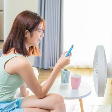 A woman sitting on a couch, looking at her mobile phone in front of a fan