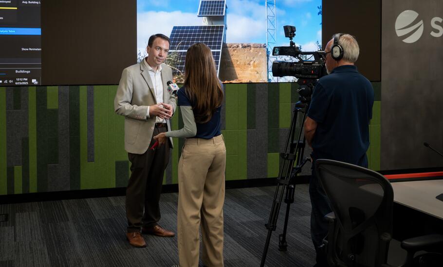 A media interview takes place at SDG&E's WCRC with illuminated screens in the background.