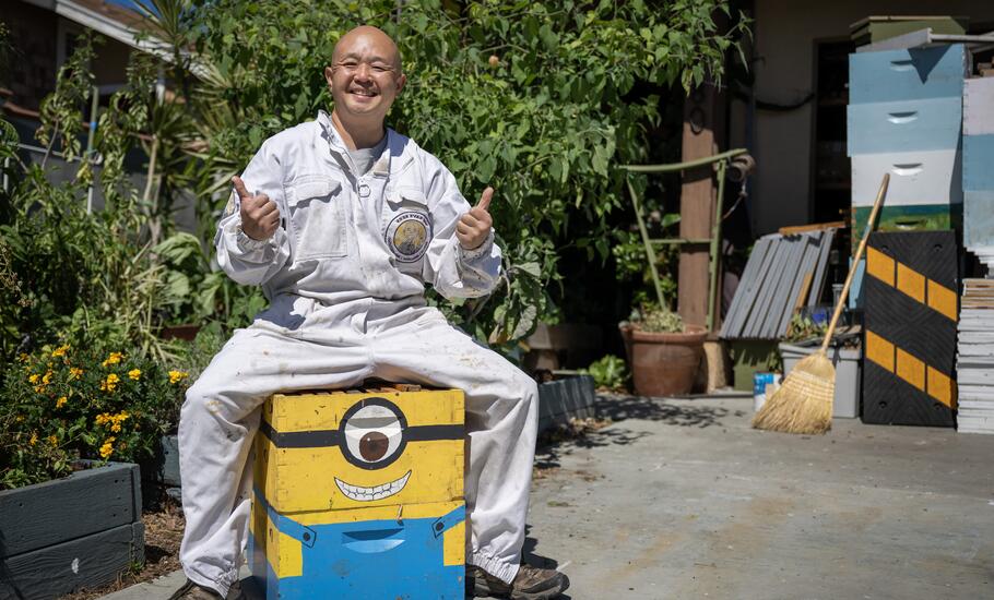 Dan the Bee Man poses for a photo, sitting on a colorful wood box and giving a thumbs up sign. 