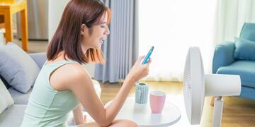 A woman sitting on a couch, looking at her mobile phone in front of a fan