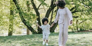 A woman holds a young childs hand, walking through a scenic setting with trees.