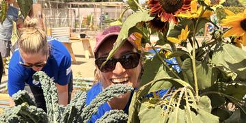 A smiling female volunteer pokes her head through garden greenery, with a vibrant sunflower positioned above her. 
